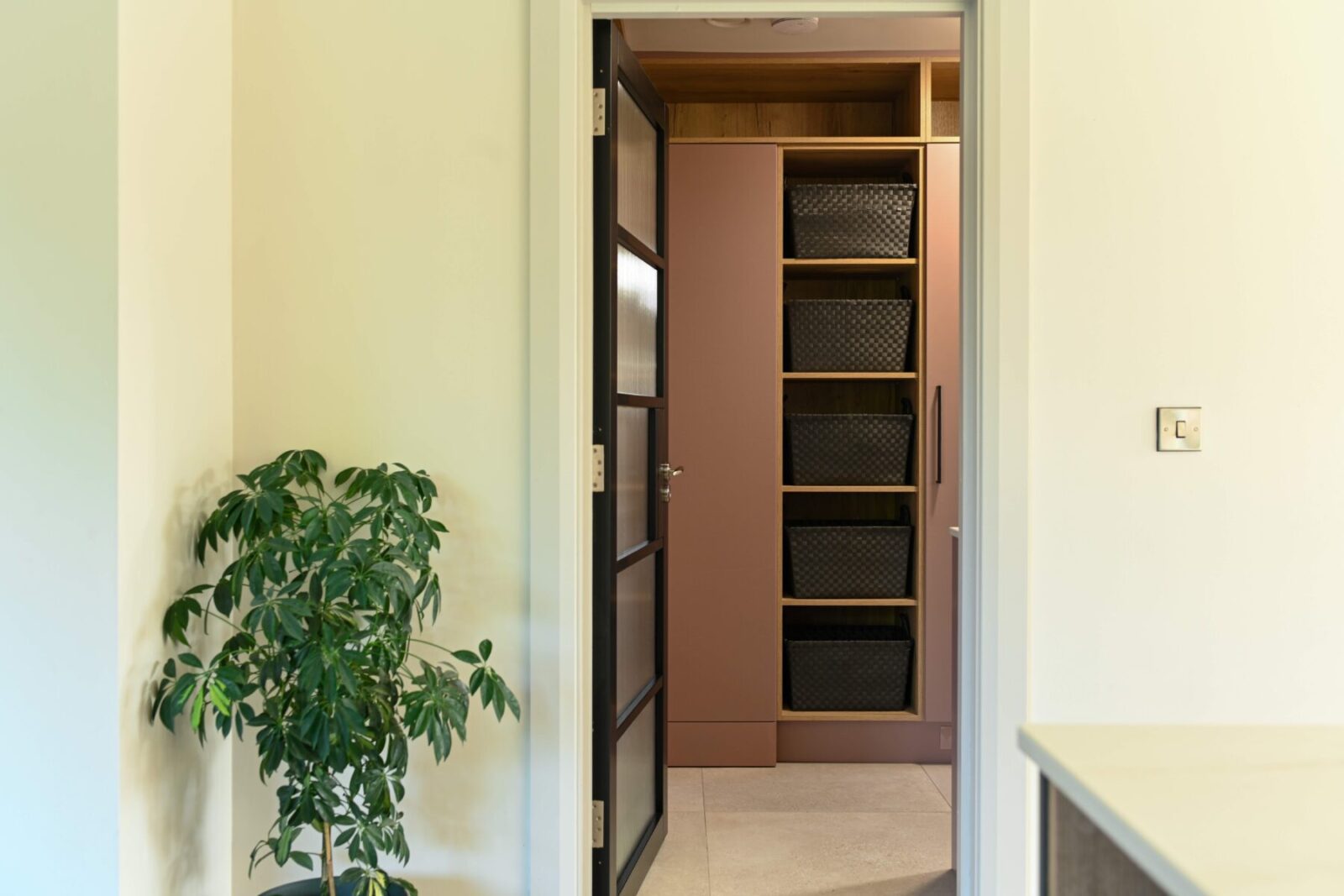 A blush pink utility room viewed through a black-framed glass door, featuring floor-to-ceiling cabinetry with woven storage baskets and natural oak shelving for a mix of concealed and open storage solutions.